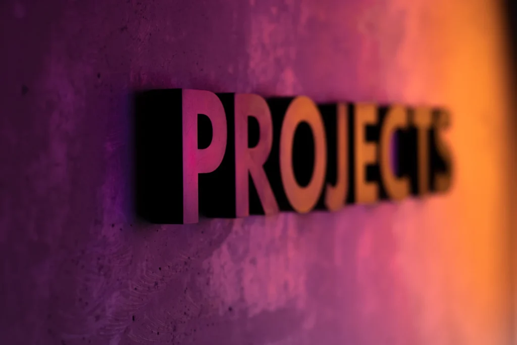 Schriftzug „Projects“ an einer Wand in pinkem und orangem Licht, Symbolbild für den Projektauftrag